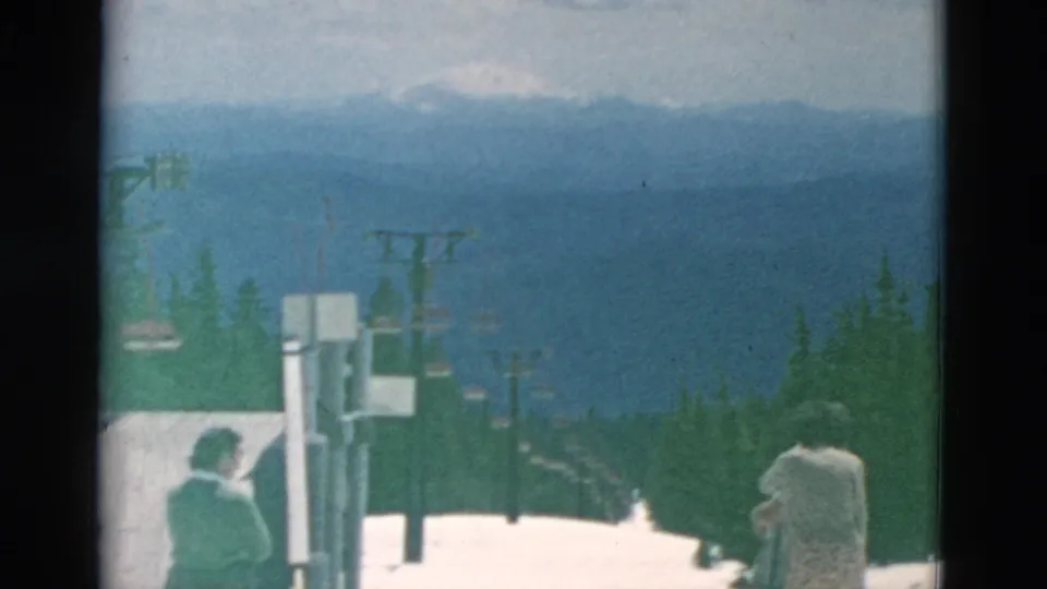 A 1958 8mm home-movie frame shows two bundled adults near a ski lift, snowbanks, evergreens, and distant mountain ridges.