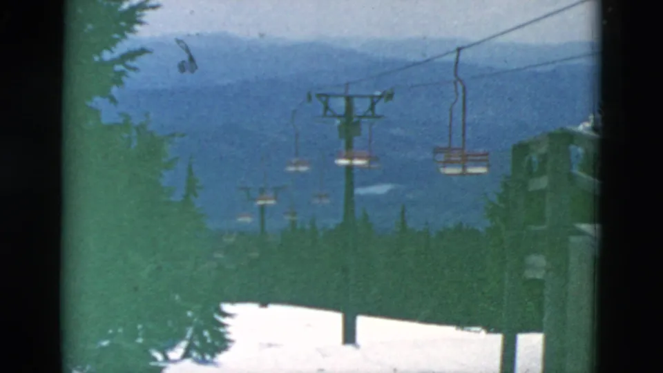 A snowy mountain ski-lift line appears above dark evergreens, with empty chairs, lift towers, and blue ridges in the background.