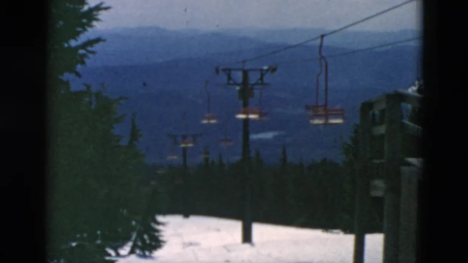 The chairlift continues across a snow-covered slope, with towers and empty chairs silhouetted against distant blue mountain scenery.