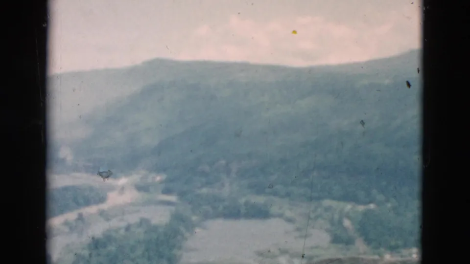 A soft, grainy 8mm view looks across a mountain valley or slope with snow patches, wooded areas, and a pale sky.