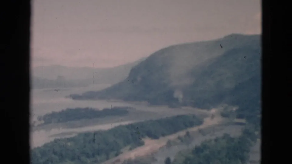 Faded 8mm still showing a soft, distant waterway bordered by trees and a rounded mountain slope.