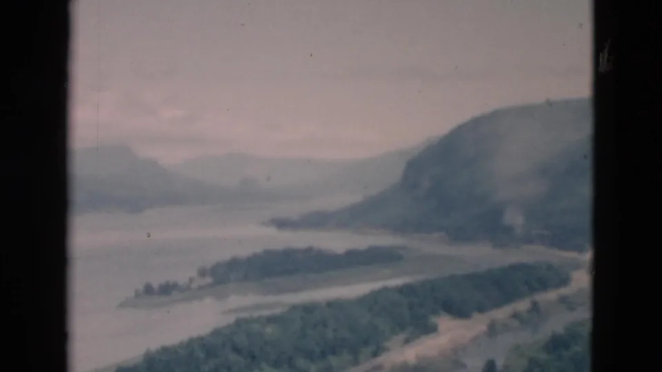 Soft color 8mm frame of a broad waterway, forested foreground, and distant mountain forms beneath an overcast sky.
