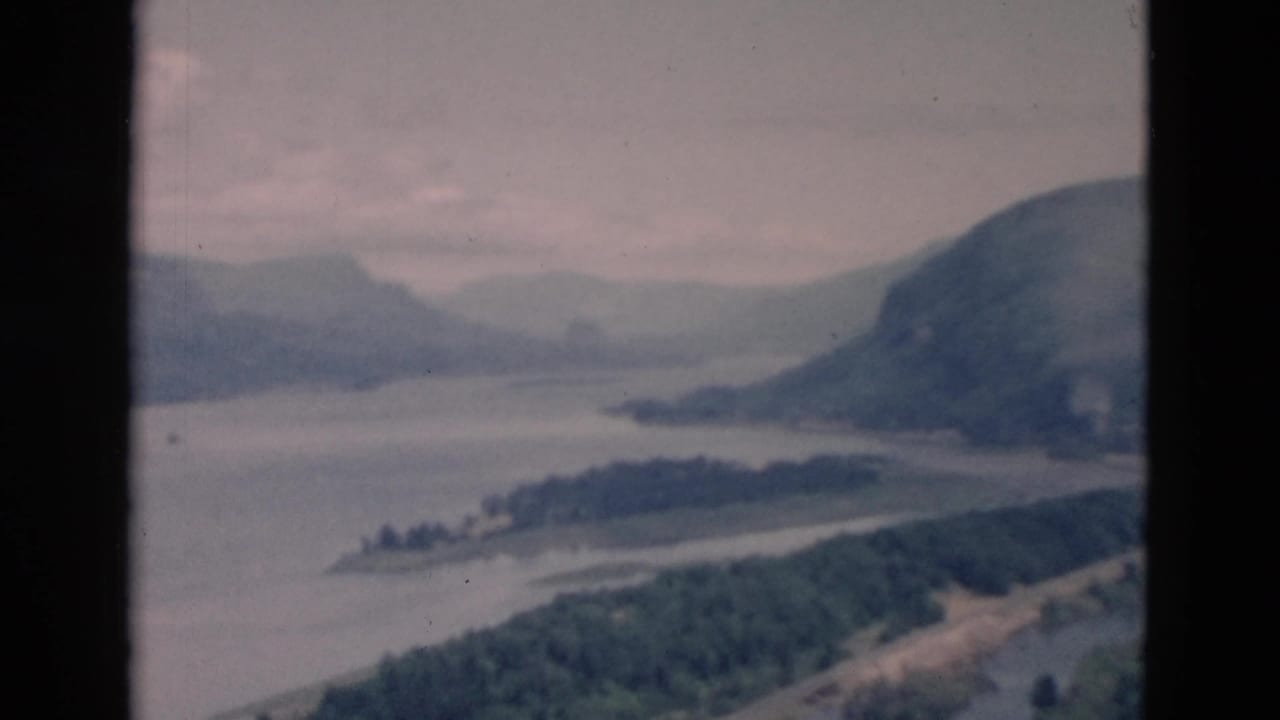 Wide river landscape with forested hills, distant mountains, and dark film borders.