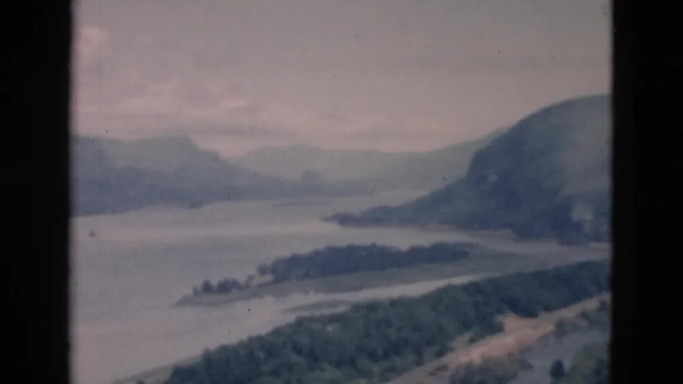 1958 8mm frame showing a broad river or lake channel between wooded hills and distant ridges under an overcast sky.