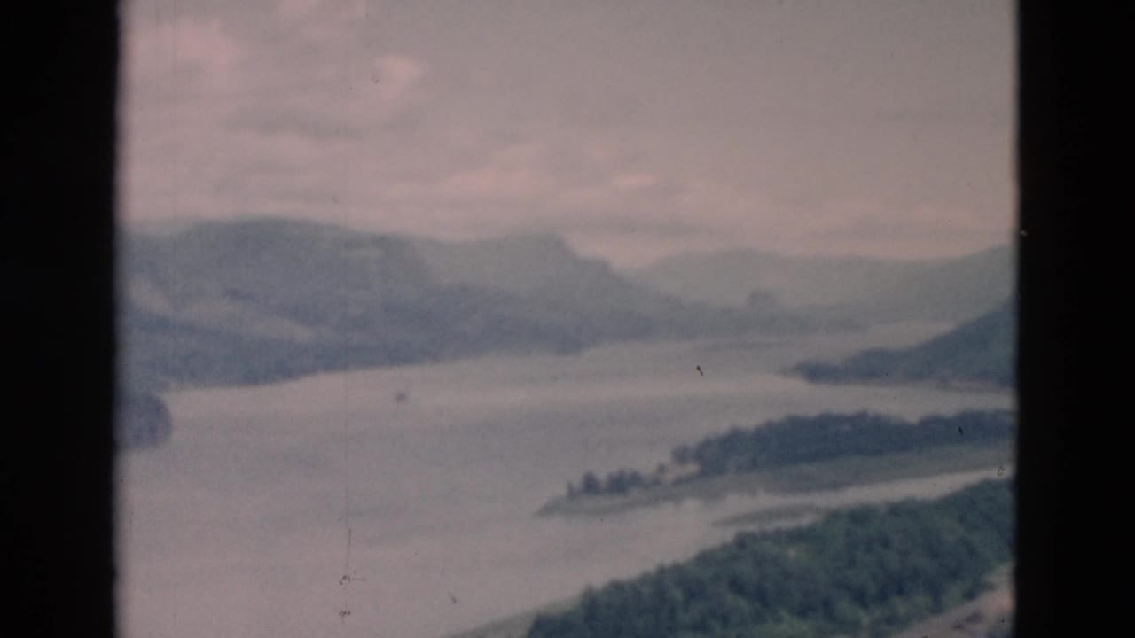 River or lake scene with low wooded land, distant mountains, and soft cloudy sky.