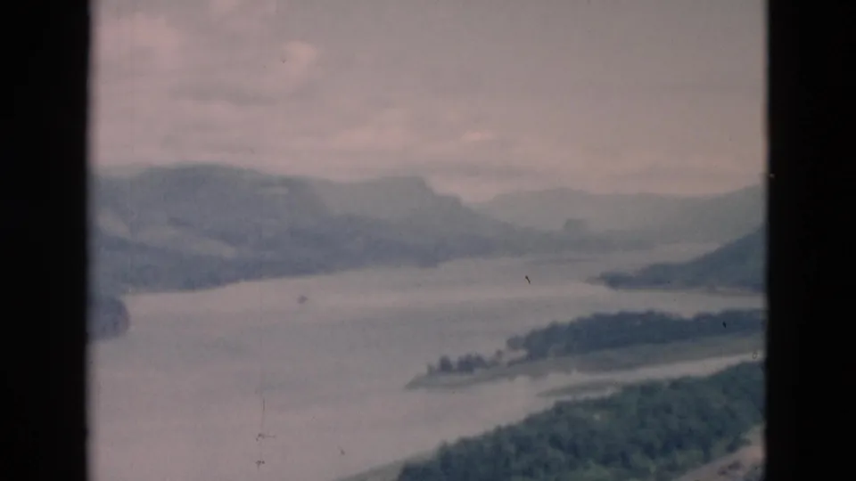 Soft 8mm view of a broad waterway, low island or peninsula, and layered mountain ridges in a likely Oregon landscape.