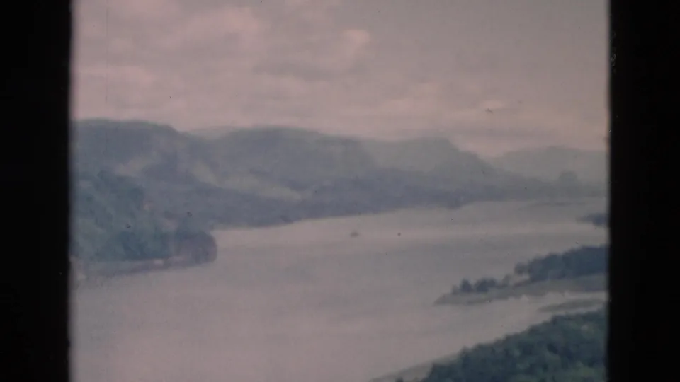 Vintage 8mm frame looking across a wide waterway toward wooded shorelines and soft distant hills, with no people or structures visible.