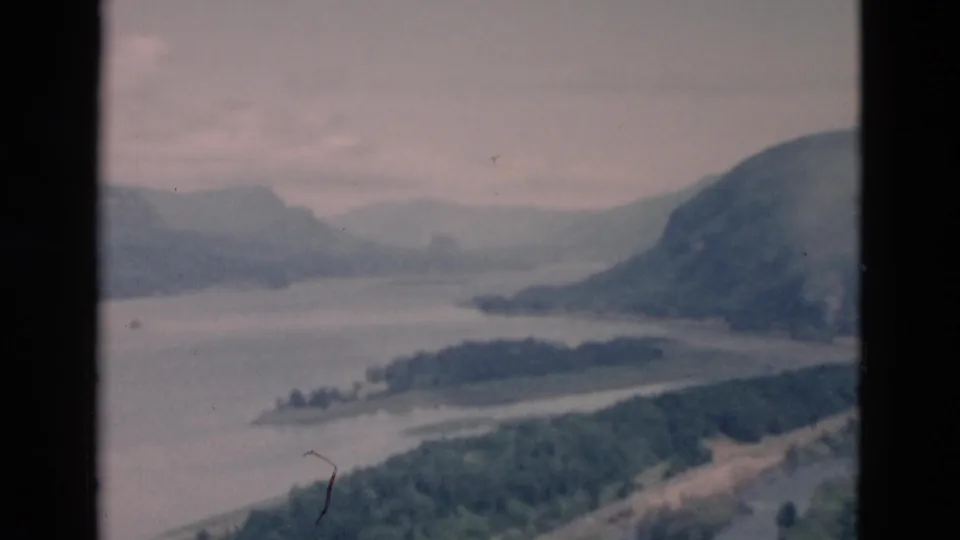 End frame of the 8mm landscape view, showing a broad river or lake, wooded point of land, and mountain slopes beneath a gray sky.