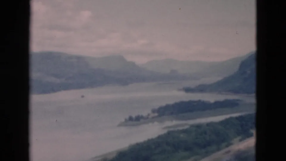 1958 8mm frame of a hazy mountain lake seen from above, with wooded slopes and soft, faded color.