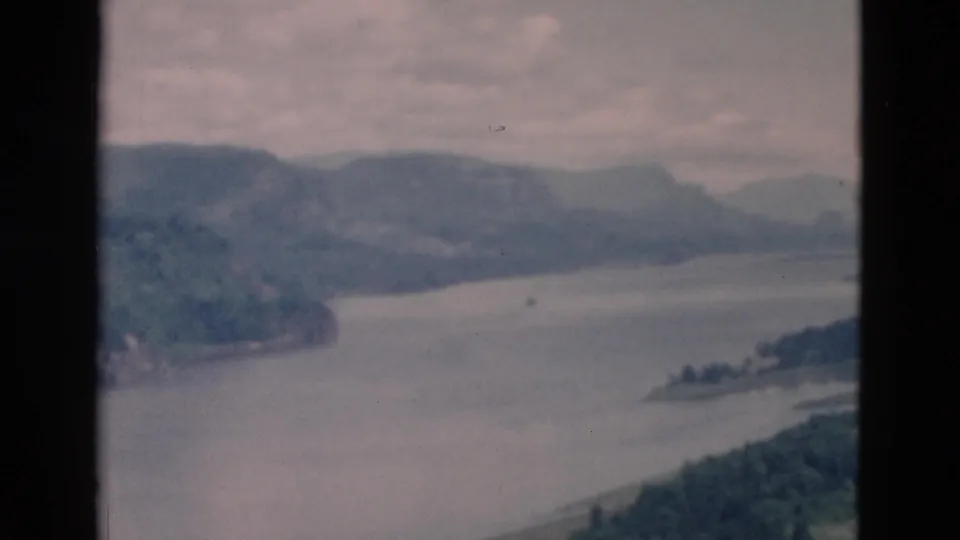 Hazy elevated view across a lake curve, forested slopes, and distant mountain ridges in soft 8mm color.