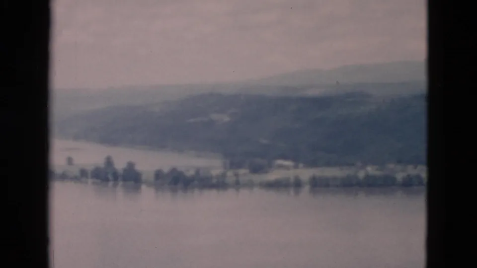 Elevated home-movie view of lake water, a tree-lined shore, and distant hills softened by haze.