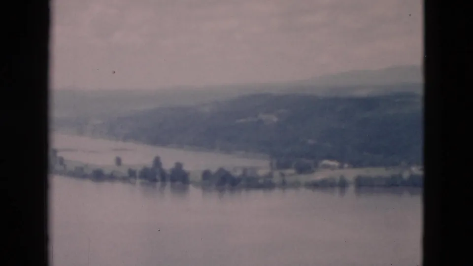 A muted 8mm frame shows calm water, a far shoreline, scattered trees, and rolling hills in soft focus.