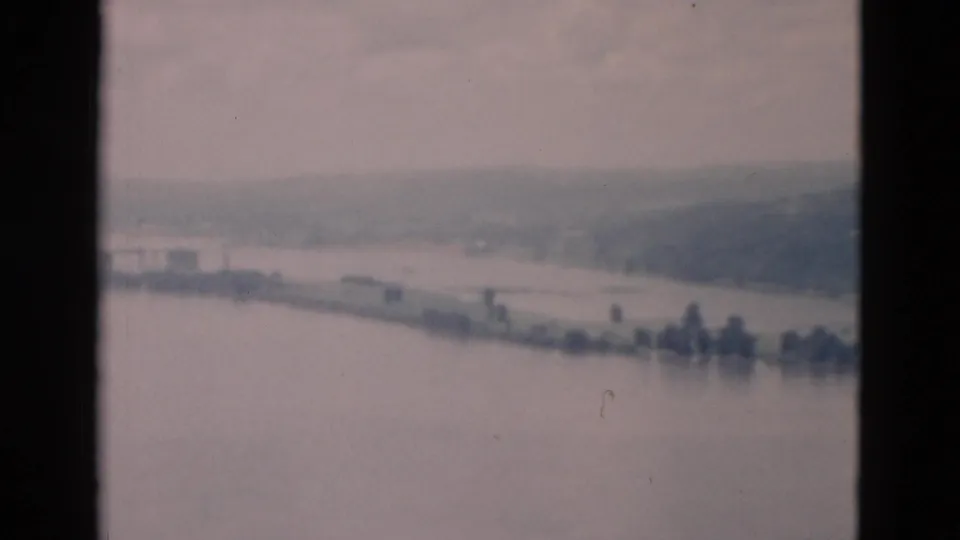 A soft, hazy 8mm frame shows open water, a thin shoreline, scattered trees, and distant hills.