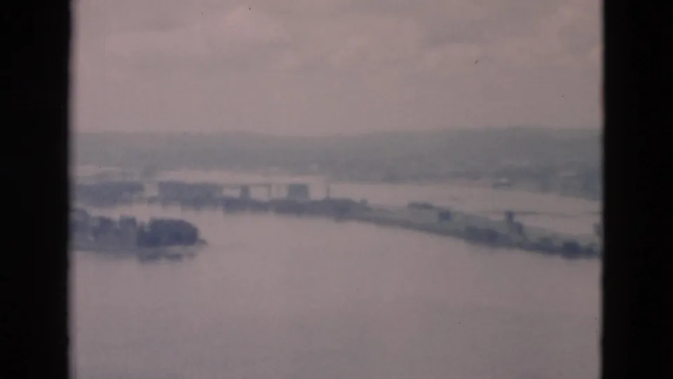 Faded 1950s 8mm frame showing a broad river, low island, hazy far bank, and faint structures from an elevated viewpoint.
