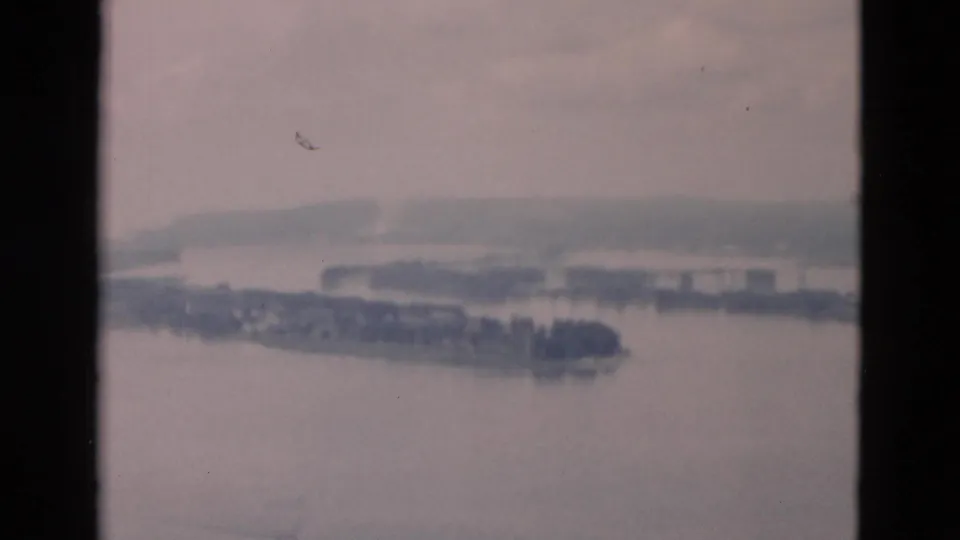Soft, faded 8mm frame of a river bend or broad channel, showing wooded island edges, far bank, and indistinct structures.