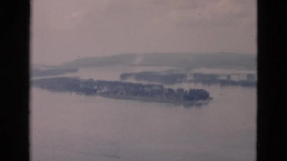 Soft, grainy 8mm frame showing a broad waterway, dark wooded landforms, and low hills under an overcast sky.