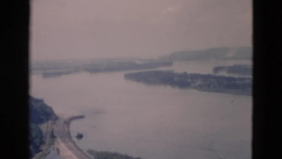 The pan reveals more shoreline from above, including water, a wooded slope, a curved road, and hazy land across the river.