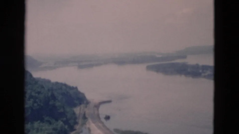 A faded 8mm frame looks over treetops and a winding road toward a broad river or bay with low, hazy hills beyond.