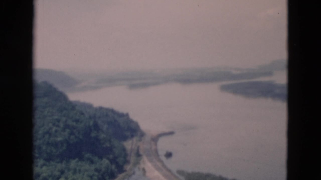 Aerial view of a lake beside forested hills with a shoreline road and dark film borders.