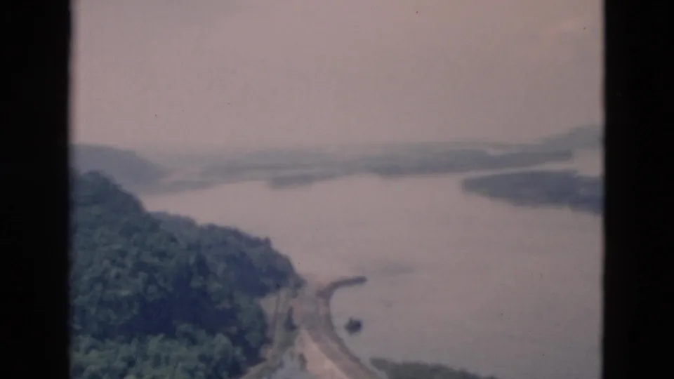 1950s 8mm-style aerial view of a hazy lake, wooded hills, and a curving shoreline road, with the exact location not confirmed.