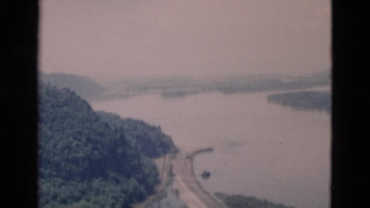 A soft 8mm aerial frame shows a lake curving around forested hills with a road along the shore and no visible people or signage.