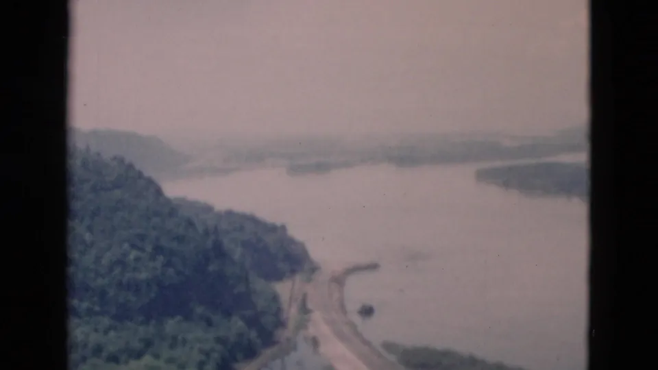 A soft 8mm aerial frame shows a lake curving around forested hills with a road along the shore and no visible people or signage.
