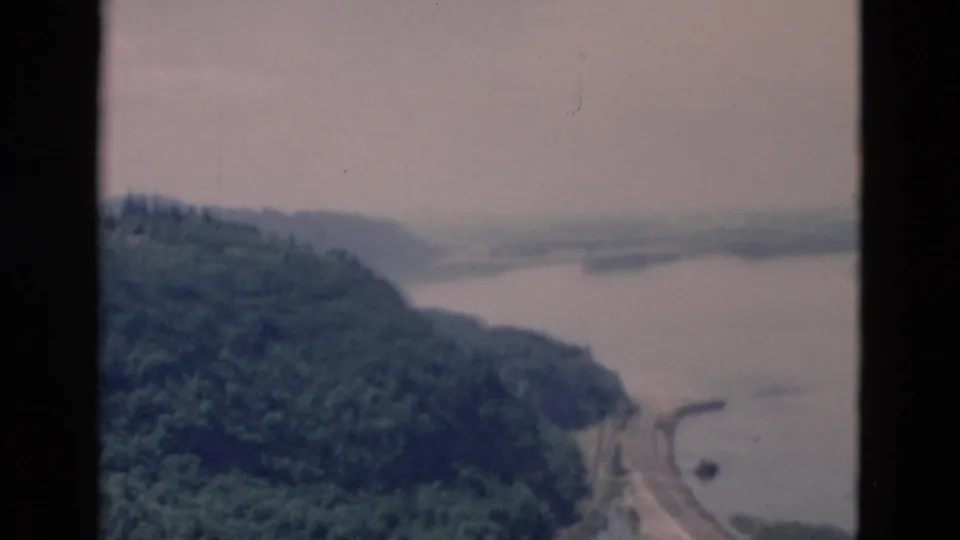 Vintage 8mm travel footage appears to look down on a forested slope, lake water, and a curving shoreline road under a pale sky.