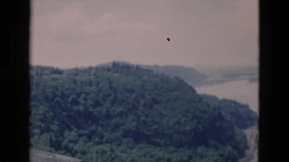 Aerial 8mm frame with a forested hill in the foreground, lake water to the right, and a hazy distant shoreline beyond.
