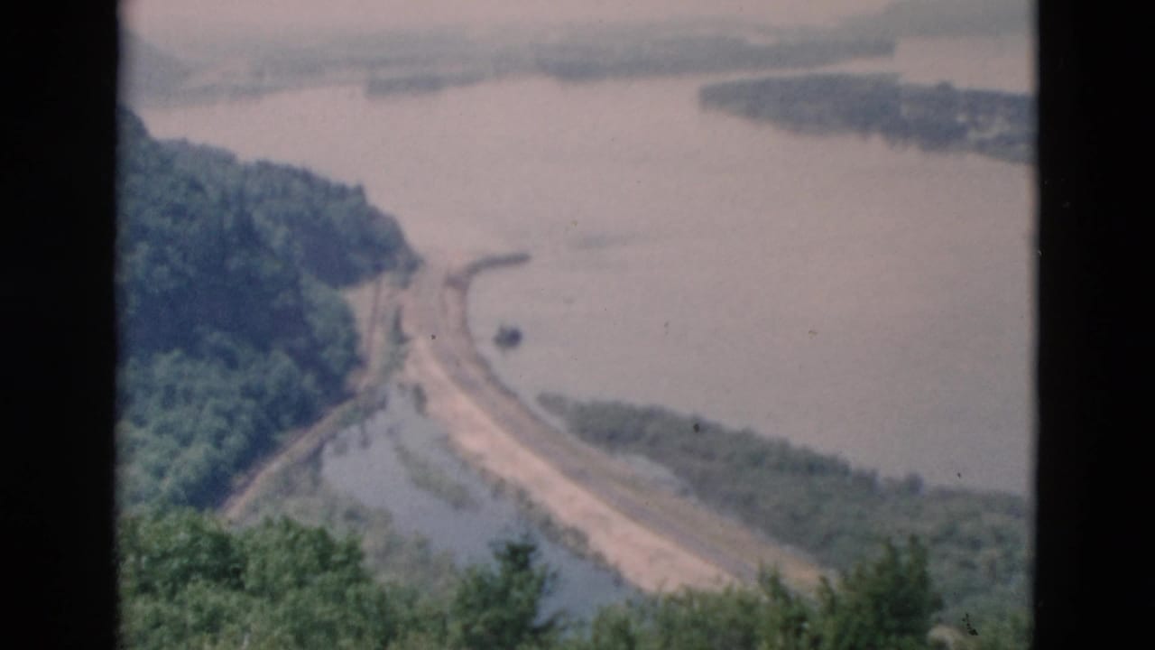 Elevated 8mm view of a river or reservoir edge with a curving road and forested slopes in the distance.