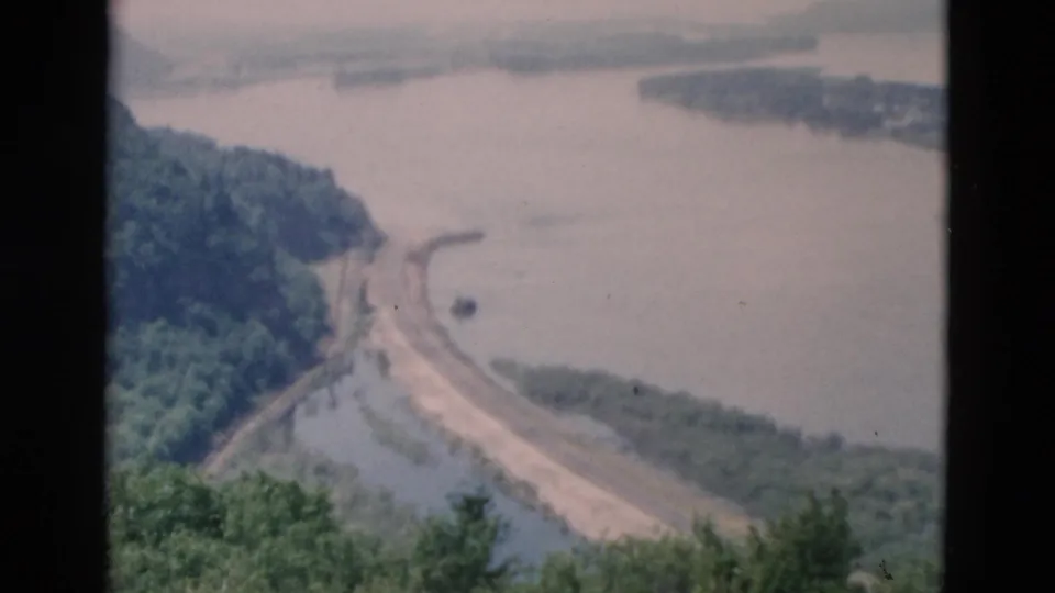 Elevated 8mm view of a river or reservoir edge with a curving road and forested slopes in the distance.