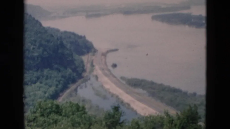 Vintage 8mm overlook shot of a road following a river or reservoir edge beneath green hills.