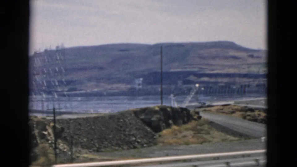 8mm view of a distant dam-like infrastructure site with a road, utility poles, earthworks, and a broad hillside.