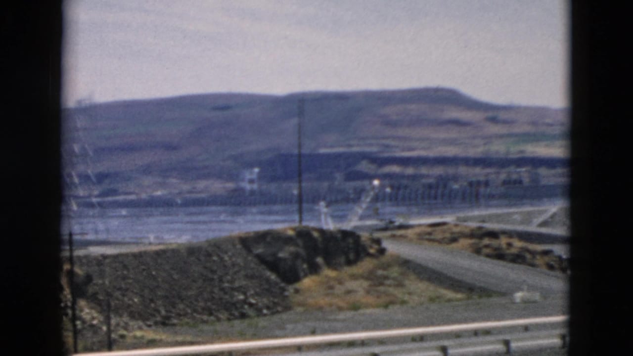 1950s 8mm roadside view toward a dam or hydroelectric facility, with guardrail, road, water, utility poles, and hazy hills.
