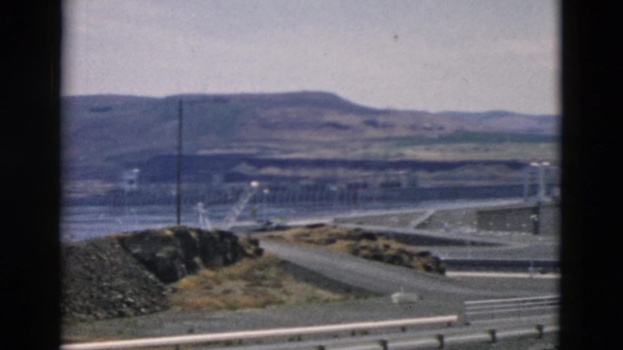 Vehicle-shot 8mm frame showing highway guardrail in front of a dam-like river facility, utility poles, water, and distant hills.