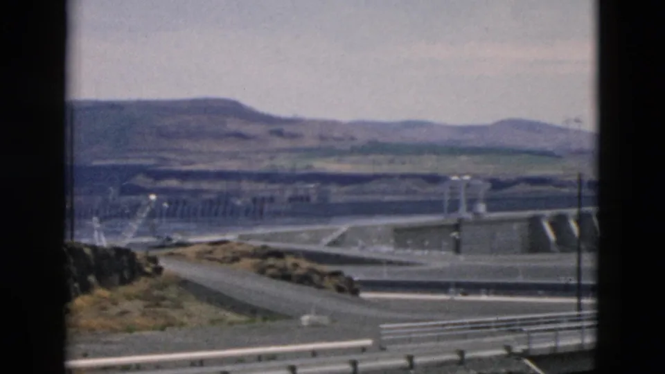 Soft 8mm frame from a moving vehicle showing road rails, a concrete dam-like structure, water, utility poles, and layered hills.