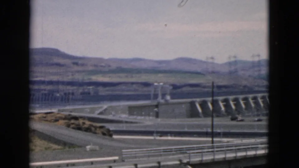 End frame of a 1950s 8mm vehicle view, with guardrails, roadway, dam-like concrete works, utility poles, and hazy hills.