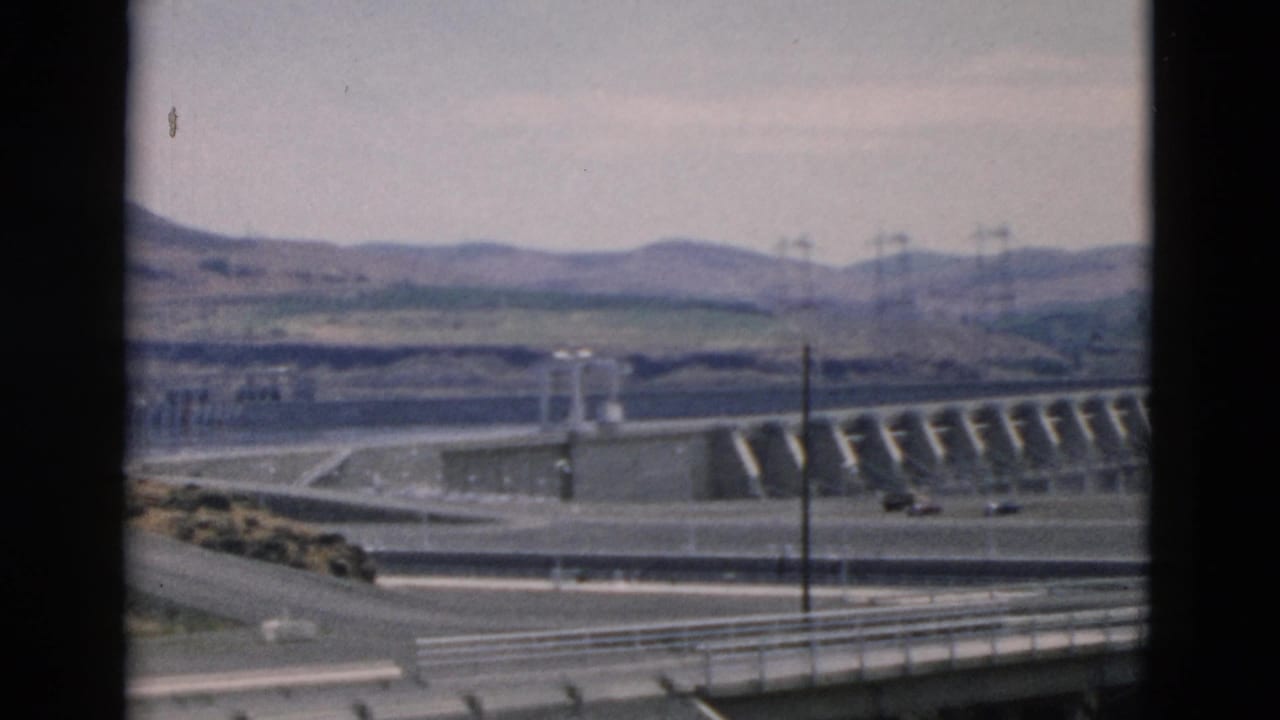 1958 8mm home-movie frame showing a roadside view toward a large dam, spillway water, utility poles, cars, and low hills in Oregon per metadata.