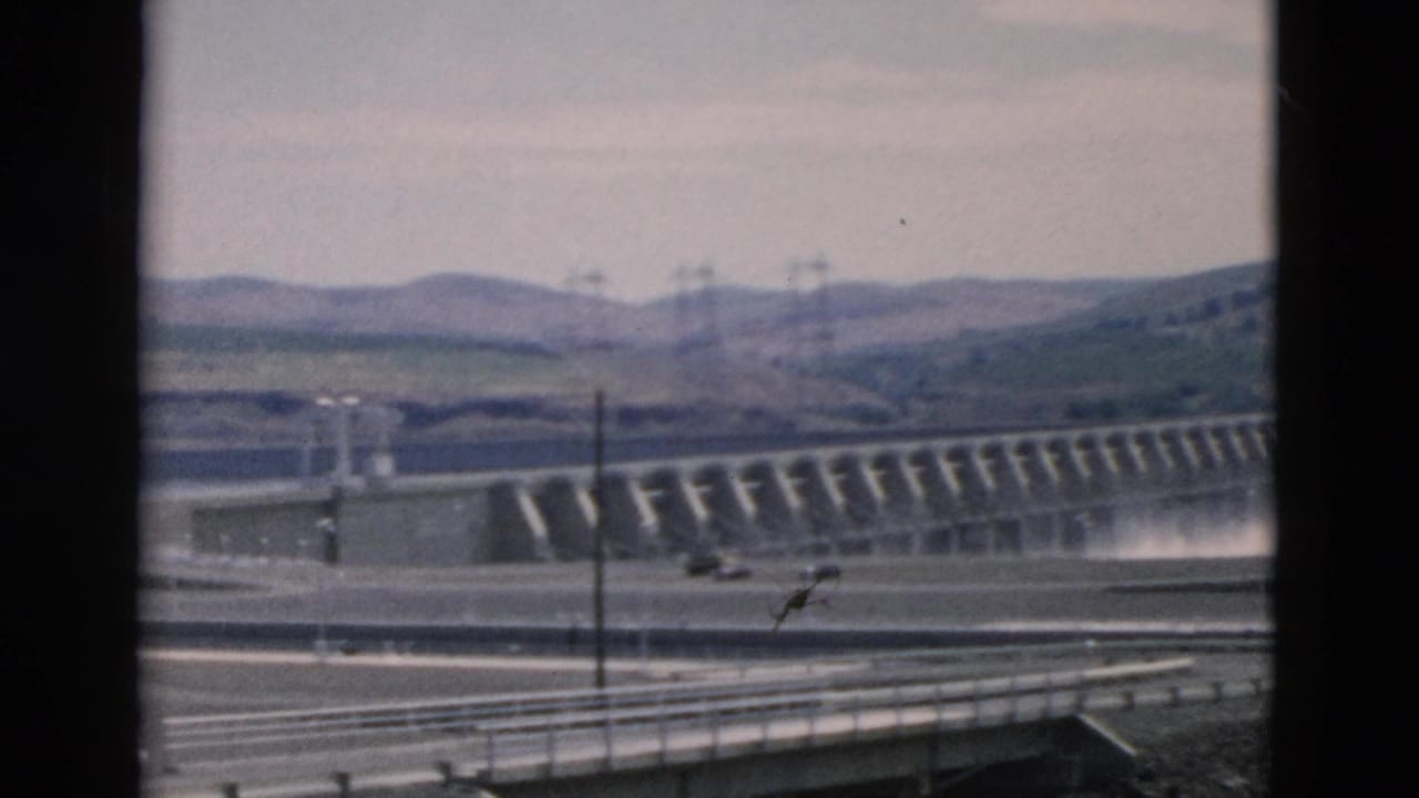 Wide Oregon dam view from 1958 home-movie footage, with water at the spillway, foreground road barriers, utility poles, and distant hills.