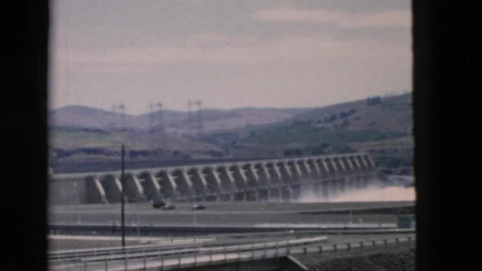 Soft 8mm frame of a long dam face and water release, seen from a road with guardrails and utility poles against open hills.