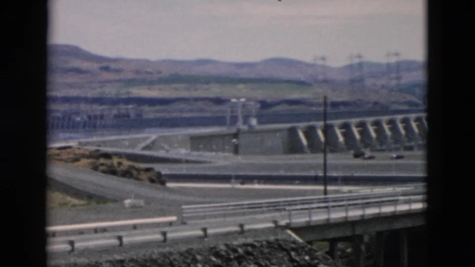 Final 1958 8mm frame showing the dam from a roadside vantage, with spillway water, road rails, utility poles, cars, and hills.