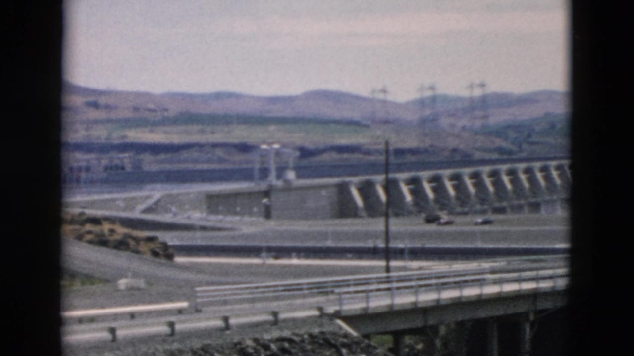 Wide view of a concrete dam with spillways, roadway, guardrails, poles, hills, and light traffic.