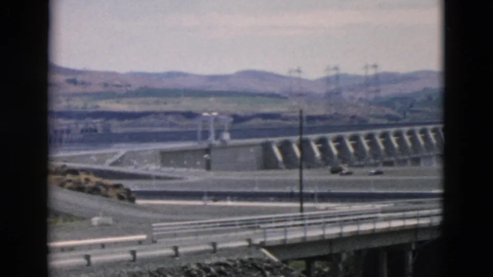 1958 8mm frame of an Oregon-attributed concrete dam scene with spillways, a foreground roadway, utility poles, distant hills, and light vehicles.