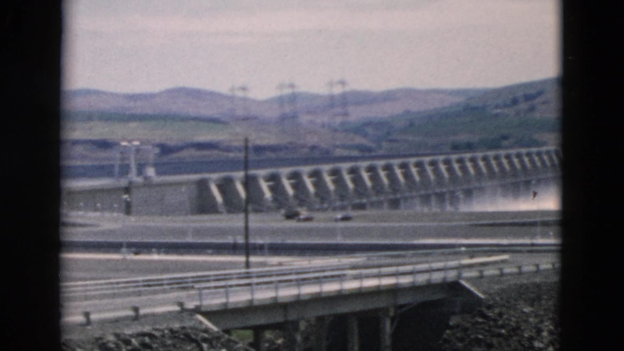 Wide 8mm view of the Oregon-attributed dam with water at the spillway, bridge or road railings in front, and power lines across the hills.