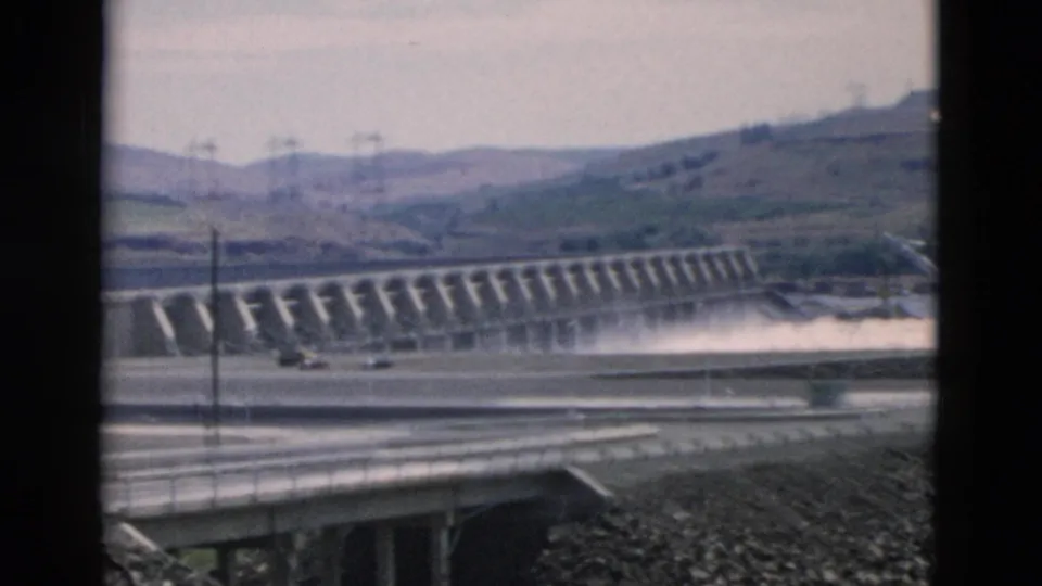 Oregon-attributed 1958 home-movie frame showing the dam from a lower roadside angle, with water spray, bridge railings, rocky ground, and hills.