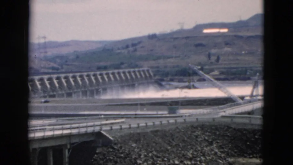 Final 8mm frame of the Oregon-attributed dam scene, with spillway water, foreground bridge, rocky ground, hillside roads, utility poles, and light flare.