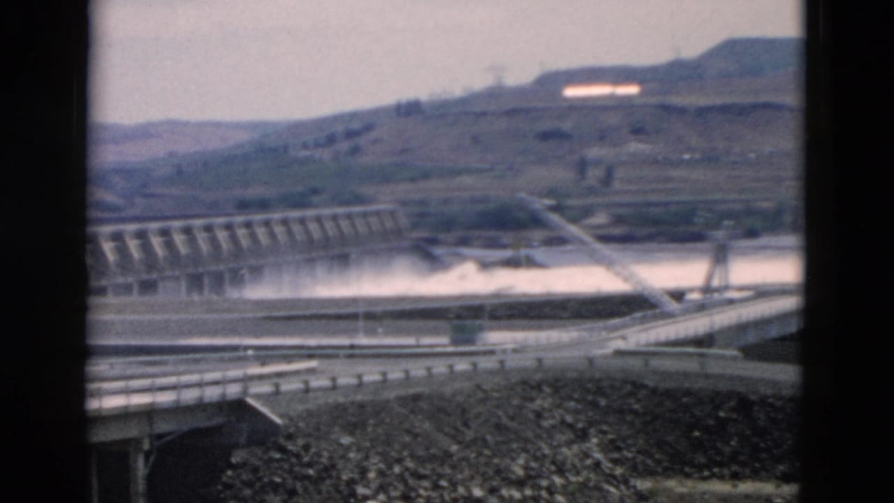 Concrete dam spillway and road approach seen across a river valley.