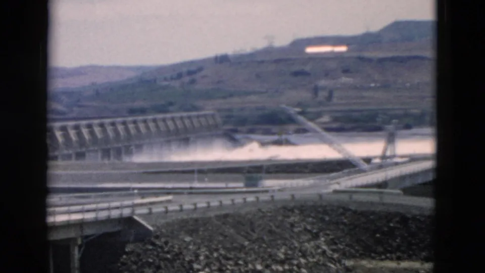 Soft 8mm frame showing a concrete dam spillway, road approach, guardrails, rocky foreground, and dry hills beyond the water.