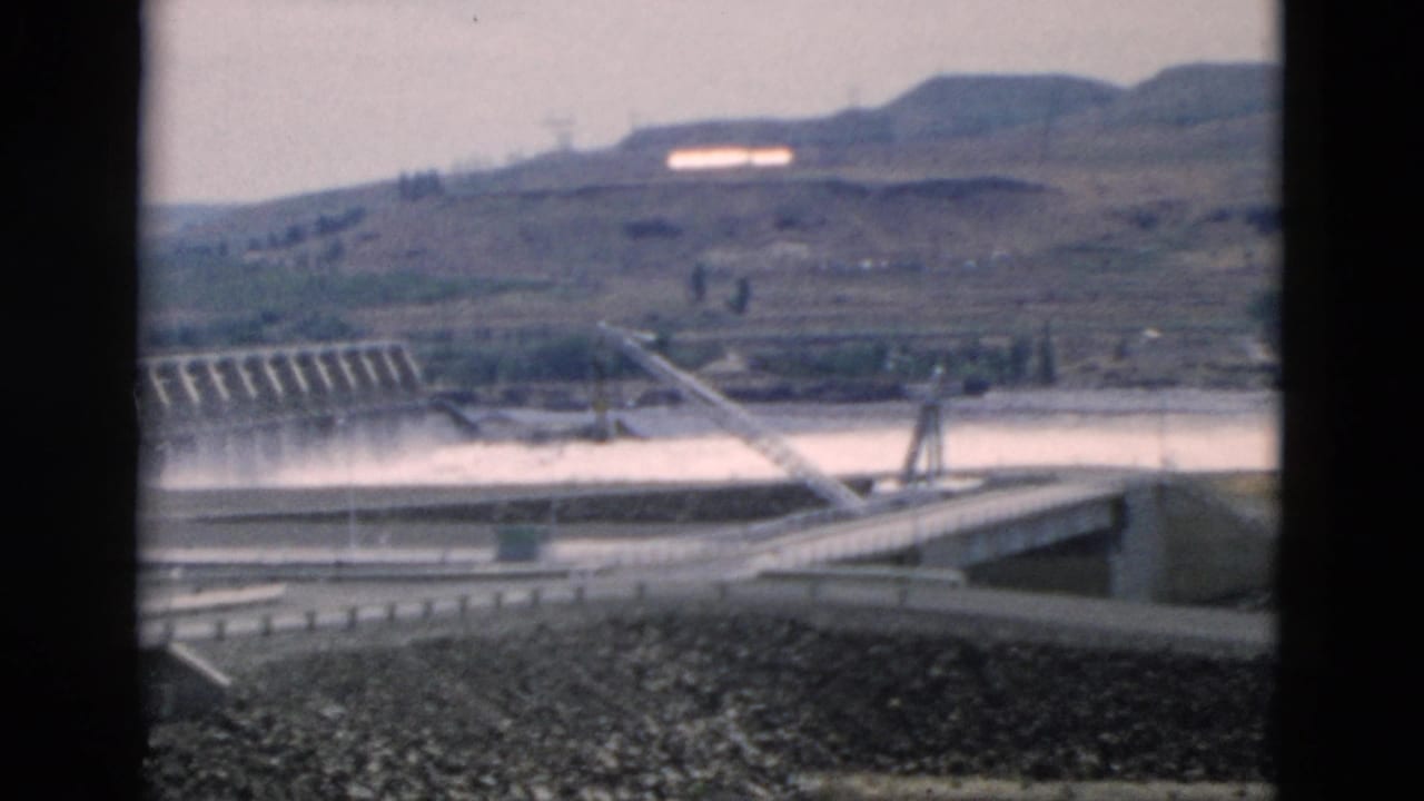 Dam, spillway water, and road bridge approach with hills in the distance.