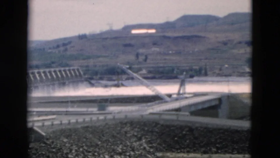 The pan holds on the dam and bridge approach, with white spillway water, paved roadways, a rocky slope, and muted hills in the background.