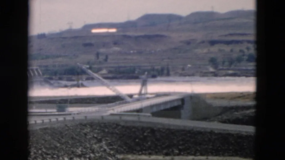 Handheld 8mm view of a dam-side road, spillway edge, broad water, rocky foreground, and pale hills during the continued pan.